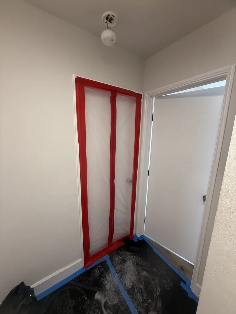 Closed plastic containment doorway with red zipper, blue tape and protected flooring.