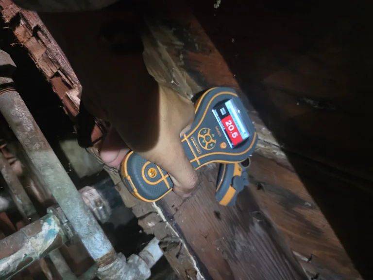 Hand holding a moisture meter against exposed framing in a dark wall cavity.