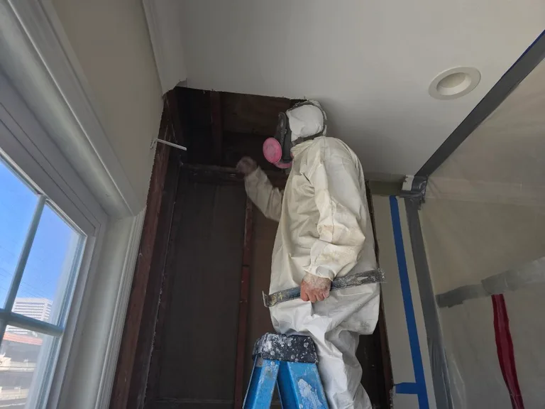 Technician in protective clothing and respirator standing on a ladder at an opened wall beside a window.