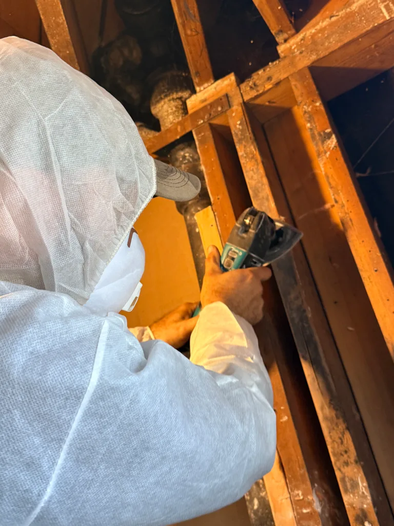 Technician in white protective clothing using a power tool on exposed wood studs.